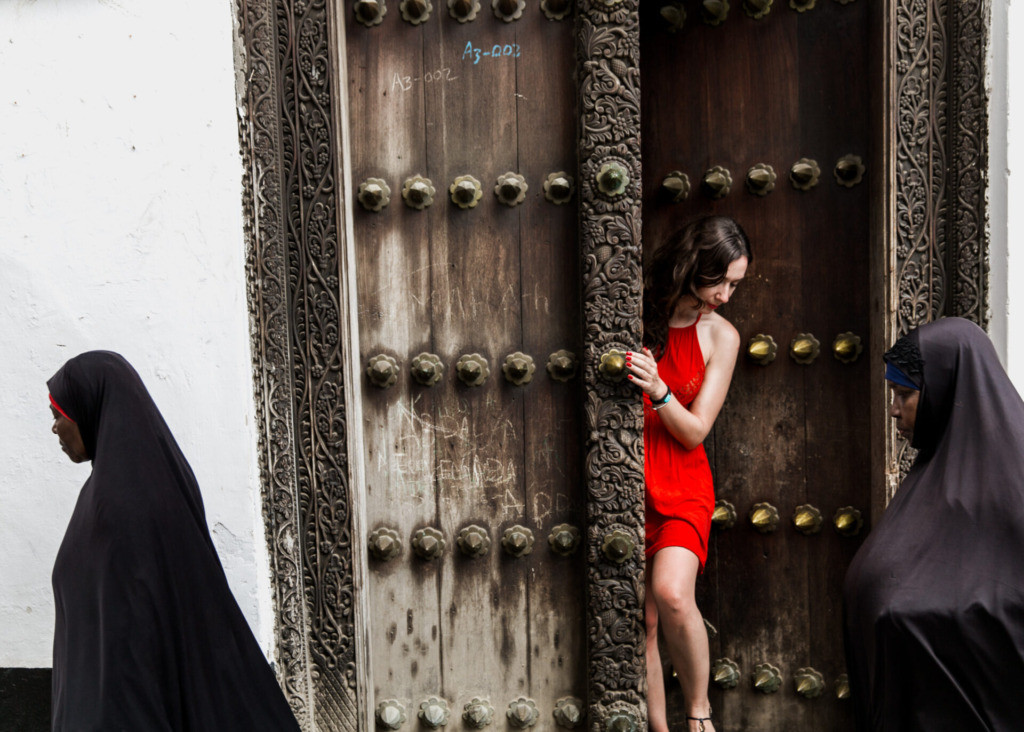 A woman in a red dress peeking out from a wooden carved door in Zanzibar.
