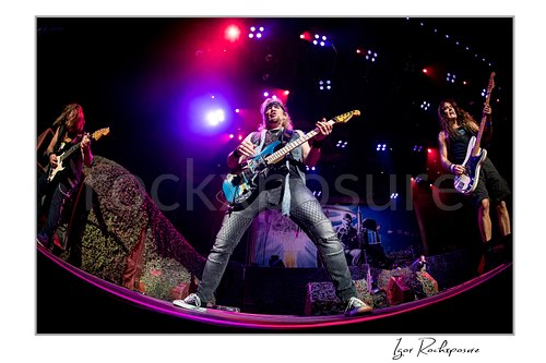 Horizontal color image of Adrian Smith, Dave Murray, and Steve Harris of Iron Maiden performing live on stage, with Smith playing guitar in the center, Murray playing guitar at left, and Harris playing bass at right under vivid purple and red concert lights.