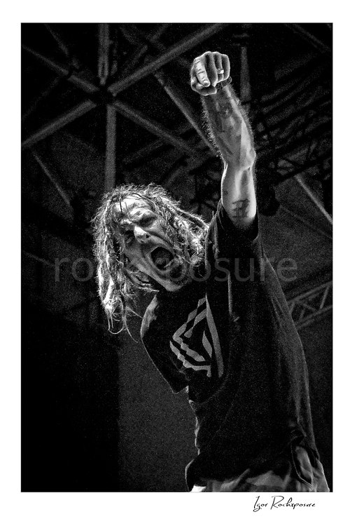 Vertical black and white image of Randy Blythe of Lamb of God with raised fist and an intense expression on stage