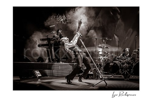 Horizontal B&W image of Depeche Mode members Peter Gordeno, Dave Gahan, and Christian Eigner performing on stage