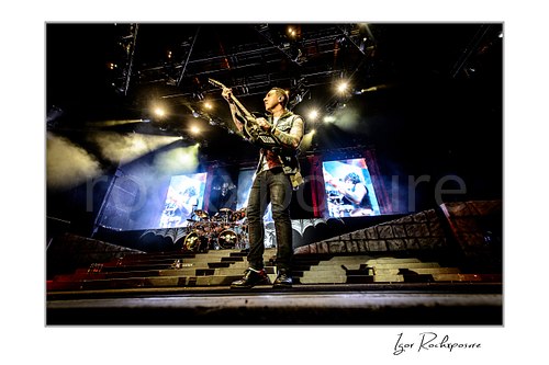 Horizontal color image of Zacky Vengeance of Avenged Sevenfold playing guitar on stage in a dramatic low-angle wide shot with bright spotlights and stage smoke