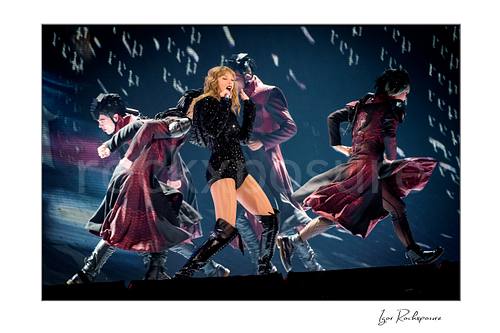 Horizontal color image of Taylor Swift singing on stage with dancers in sweeping coats under dramatic concert lighting
