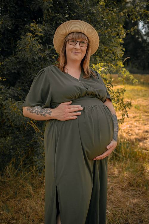 Eine schwangere Frau mit Brille, Strohhut und Tätowierungen an den Armen steht lächelnd auf einer Wiese und hält schützend ihren Babybauch. Sie trägt ein olivgrünes Maxikleid.