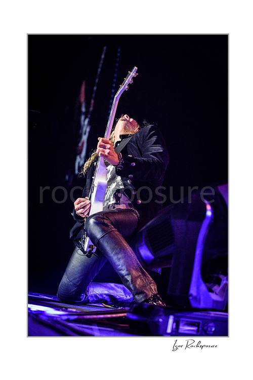 Vertical color low-angle image of Joel Hoekstra of Whitesnake performing live on stage, kneeling while playing electric guitar under purple concert lighting