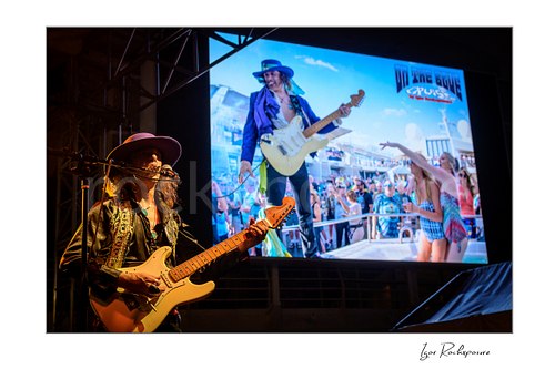Horizontal color image of Randy Hansen playing guitar on an outdoor ship deck stage with a large screen behind him showing another live performance image