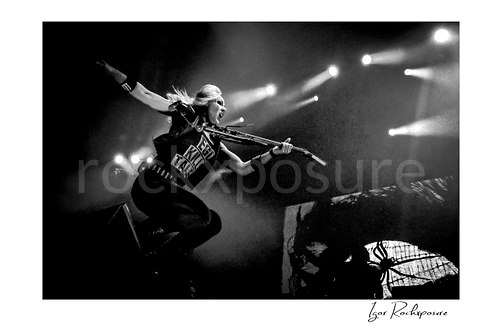 Horizontal black and white image of Nita Strauss performing live on stage with an electric guitar, captured in midair with dramatic motion