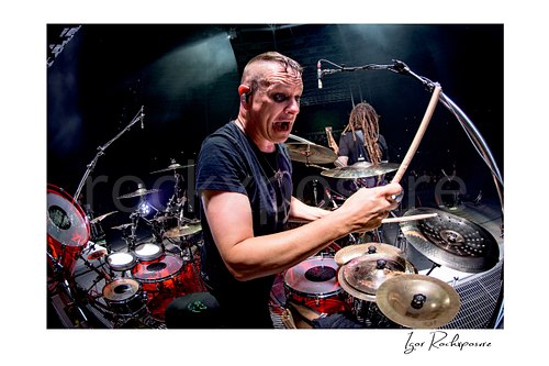 Horizontal color image of Korn drummer Ray Luzier at the drums with a drumstick raised, a scary expression, and bright stage lighting