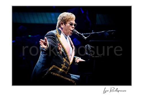 Horizontal color image of Elton John singing at the piano with one hand extended in a dark blue concert scene