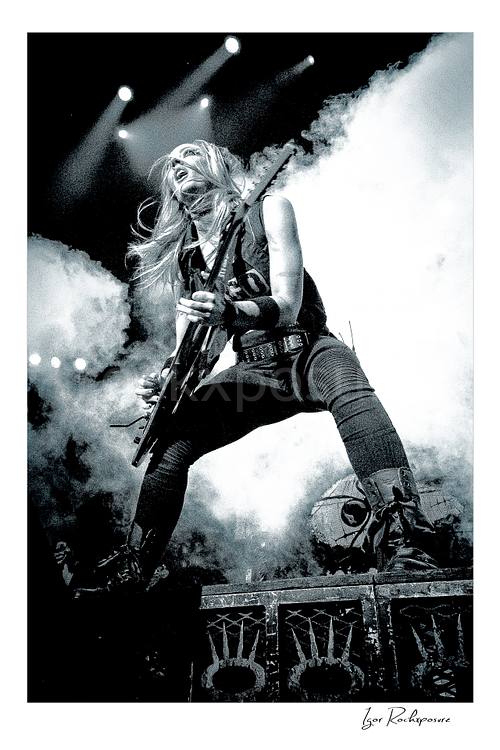 Vertical black and white image of Nita Strauss performing live on stage with an electric guitar, surrounded by thick stage smoke