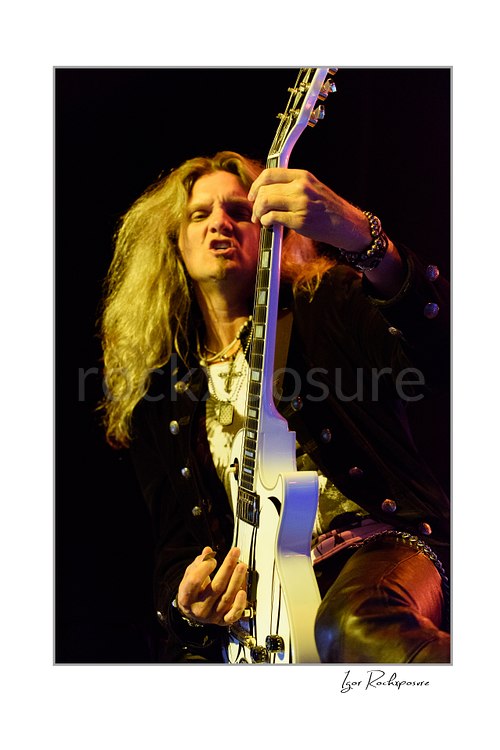 Vertical color close-up image of Joel Hoekstra of Whitesnake performing live on stage, playing white electric guitar under warm concert lighting