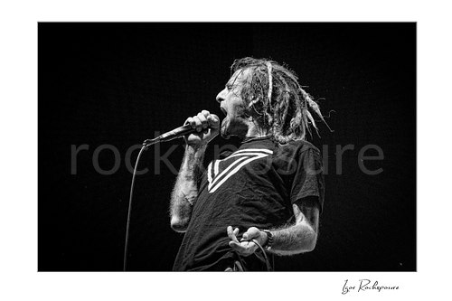 Horizontal black and white image of Randy Blythe of Lamb of God singing into a handheld microphone in side profile with dreadlocks