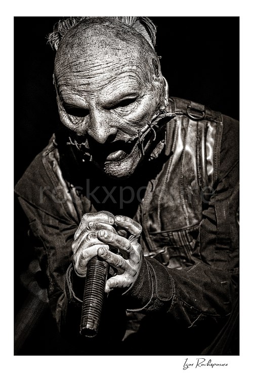 Vertical black and white image of Corey Taylor of Slipknot in a tight masked close-up gripping a handheld microphone with both hands