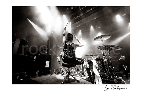 Horizontal black and white image of Zoltan Chaney playing drums from behind on stage under bright concert lights