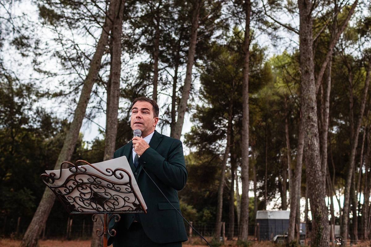 Le p&egrave;re de la mari&eacute;e en costume vert, se tenant devant un lutrin &eacute;l&eacute;gant, s'adresse aux invit&eacute;s avec un micro, dans un cadre ext&eacute;rieur entour&eacute; de pins, offrant des mots lors de la r&eacute;ception du mariage &agrave; N&icirc;mes
