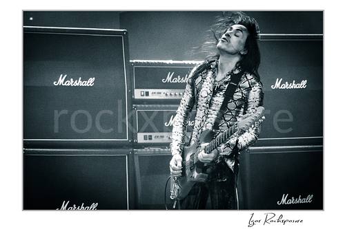 Horizontal black and white image of Nuno Bettencourt performing live on stage with an electric guitar, windblown hair, and a wall of Marshall amplifiers behind him