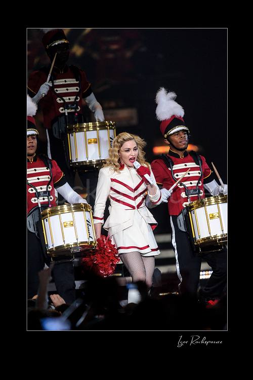 Vertical color image of Madonna singing into a microphone in a white and red stage outfit while marching drummers perform around her