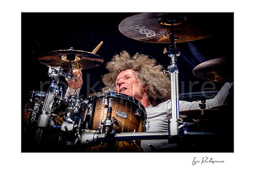 Horizontal color image of Tommy Aldridge of Whitesnake performing live on stage behind the drum kit, captured in a close-up view between cymbals and drums