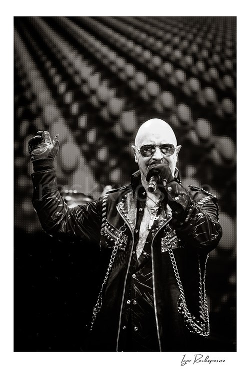 Vertical black and white image of Rob Halford of Judas Priest performing live on stage, singing into a microphone while wearing sunglasses and a studded leather coat