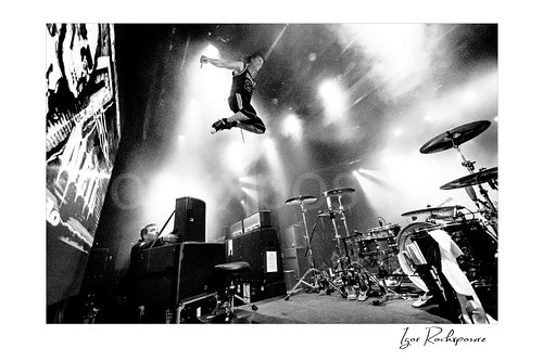 Horizontal black and white image of Zoltan Chaney leaping high on stage beside a drum kit under bright concert lights