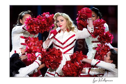 Horizontal color image of Madonna performing with dancers and bright red pom-poms in a theatrical live stage scene