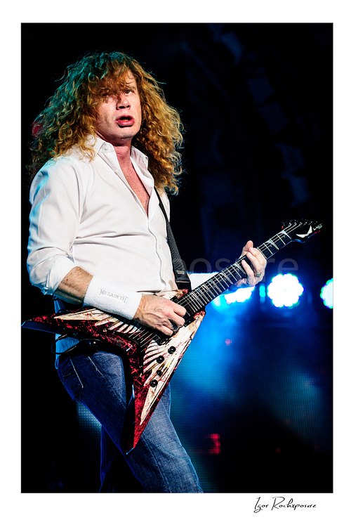 Vertical color image of Dave Mustaine of Megadeth playing guitar on stage with curly hair and blue stage lights