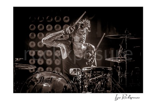 Horizontal black and white image of Tommy Lee performing at the drum kit during a Mötley Crüe concert, holding raised drumsticks under dramatic stage light