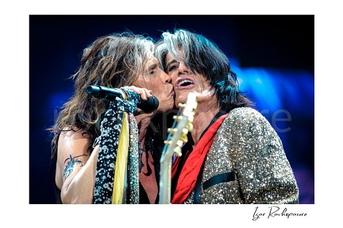 Horizontal color image of Steven Tyler and Joe Perry of Aerosmith performing live on stage in a close-up moment with a handheld microphone, scarf-wrapped mic stand, glitter jacket, and red scarf