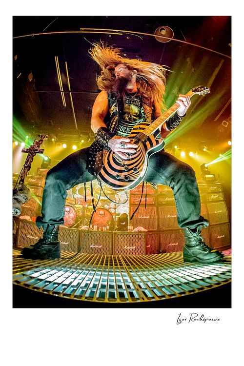 High-energy vertical color concert photography of Zakk Wylde in his signature wide stance on a stage riser with flying hair while playing his bullseye guitar under dramatic golden stage lighting with a wall of amplifiers and speakers in the background