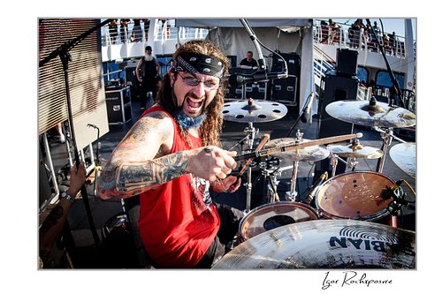 Horizontal color image of Mike Portnoy at the drum kit holding crossed drumsticks toward the camera with an intense expression in bright daylight