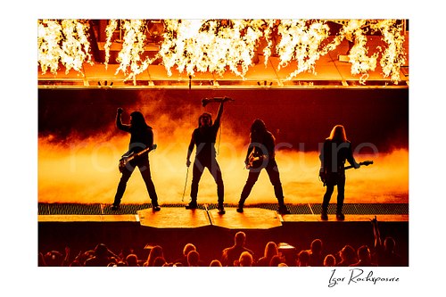 Horizontal color image of Trans Siberian Orchestra performing in silhouette with guitars, raised microphone stand, flames, and stage smoke