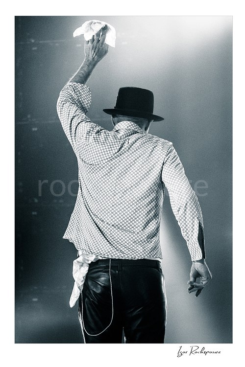Vertical black and white image of Gord Downie of The Tragically Hip seen from behind on stage, raising a white cloth in a final farewell gesture under soft concert light