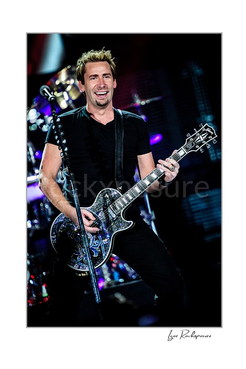 Vertical color image of Chad Kroeger of Nickelback playing guitar onstage beside a microphone stand with the drummer visible behind him
