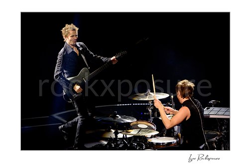Horizontal color image of Matt Bellamy and Dominic Howard of Muse performing live on stage with guitar and drums against a dark concert background