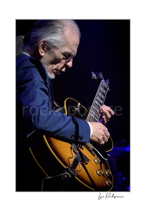 Vertical color image of Steve Howe of Yes playing guitar in a tight live performance close-up under cool blue concert light