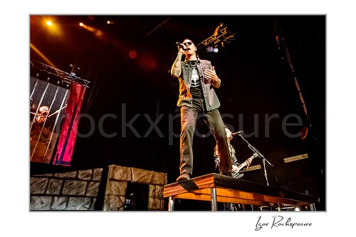 Horizontal color image of M. Shadows of Avenged Sevenfold singing on a stage riser with a microphone under warm concert lighting