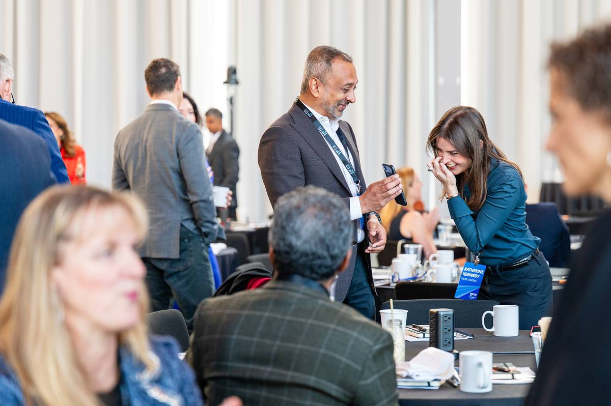 A man holds up a phone as a woman laughs, surrounded by other attendees at an executive summit in New York City.