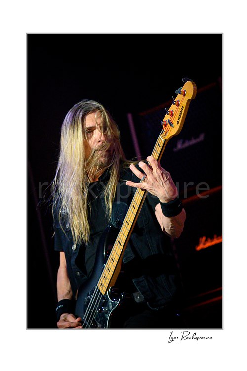 James LoMenzo of Megadeth watches his hand on an upright bass neck, with fingers spread across the fretboard and long blond hair over his vest.