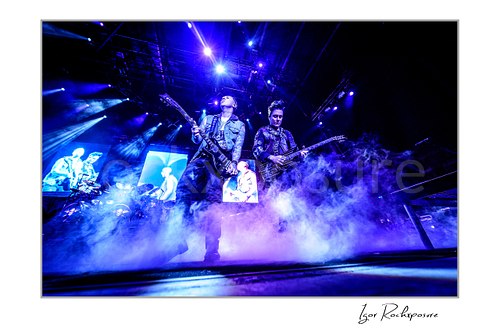Horizontal color image of Synyster Gates and Zacky Vengeance of Avenged Sevenfold performing onstage through thick blue-lit stage smoke.