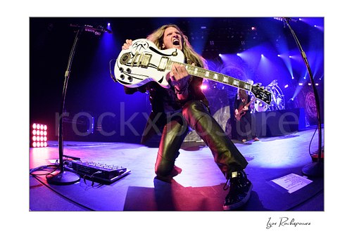 Horizontal color image of Joel Hoekstra of Whitesnake performing live on stage in a dramatic low-angle shot, kneeling with his white guitar under vivid purple concert lighting