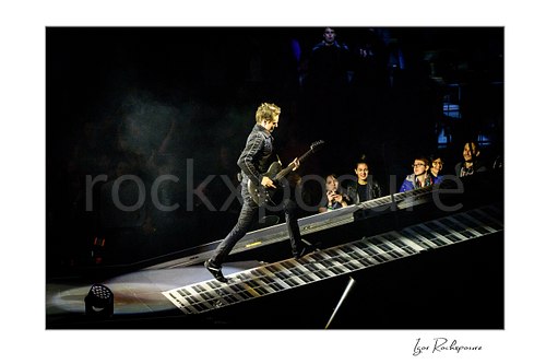 Horizontal color image of Matt Bellamy of Muse walking up a piano-key stage ramp with guitar under dramatic concert lighting