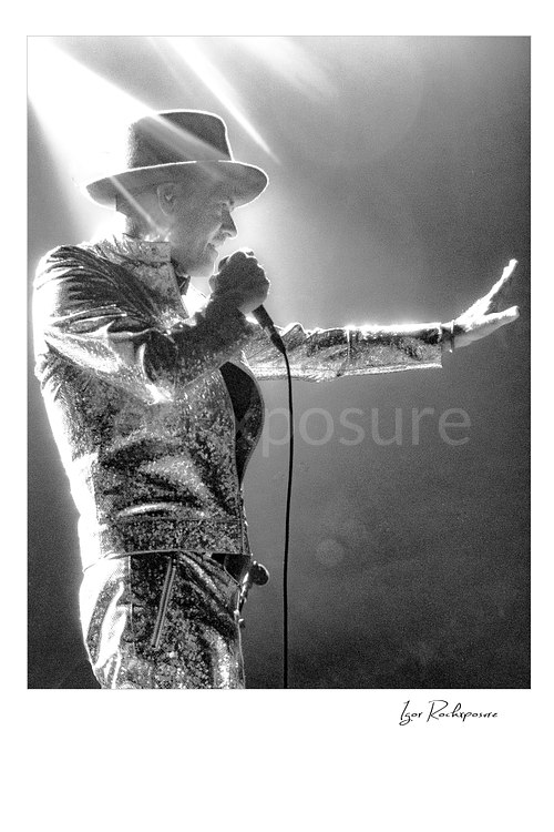 Vertical black and white image of Gord Downie of The Tragically Hip singing into a microphone in profile, wearing a glittering stage suit and hat with one hand extended into dramatic backlight