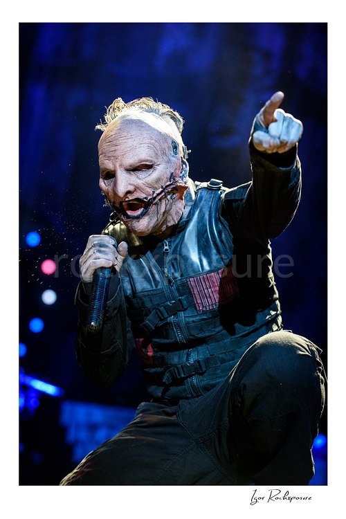 Vertical color image of Corey Taylor of Slipknot wearing a mask and holding a handheld microphone while pointing toward the crowd under blue stage light