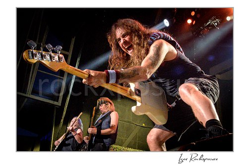 Steve Harris of Iron Maiden performing live on stage, playing bass guitar in a dramatic low-angle concert shot with other band members visible in the background