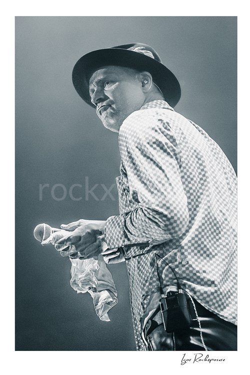 Vertical black and white image of Gord Downie of The Tragically Hip holding a microphone and white cloth in a quiet live performance moment, wearing a hat and patterned shirt under soft stage light