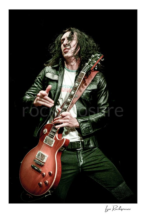 Vertical color image of Brent Woods playing guitar on stage wearing a leather jacket in a mid-performance moment