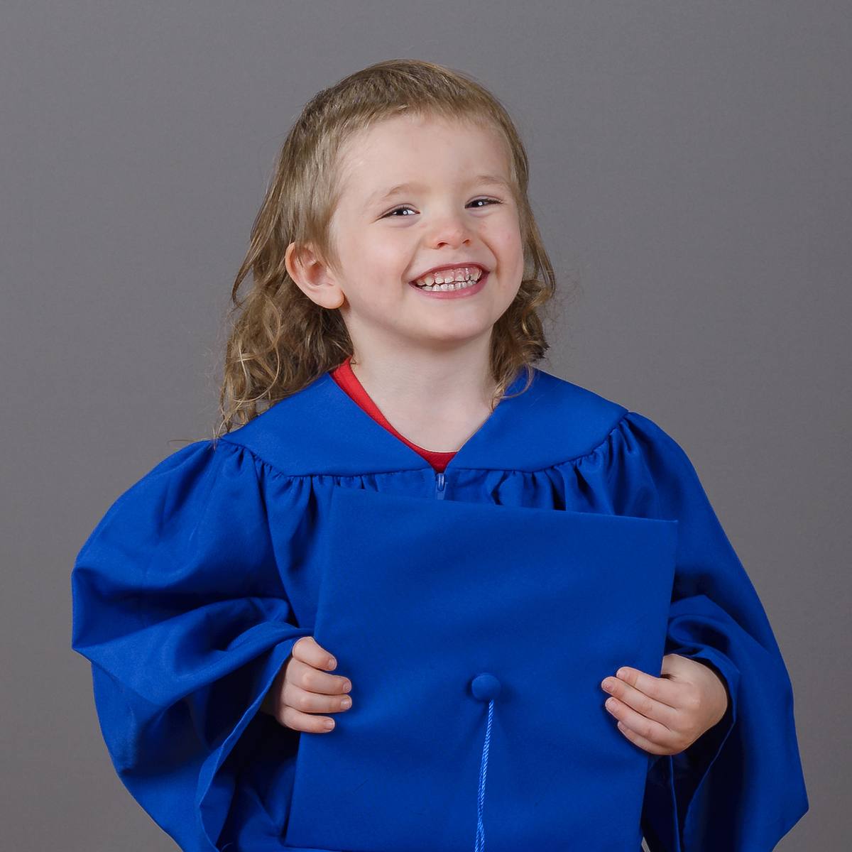 Preschool graduate in blue cap and gown holding cap on gray background
