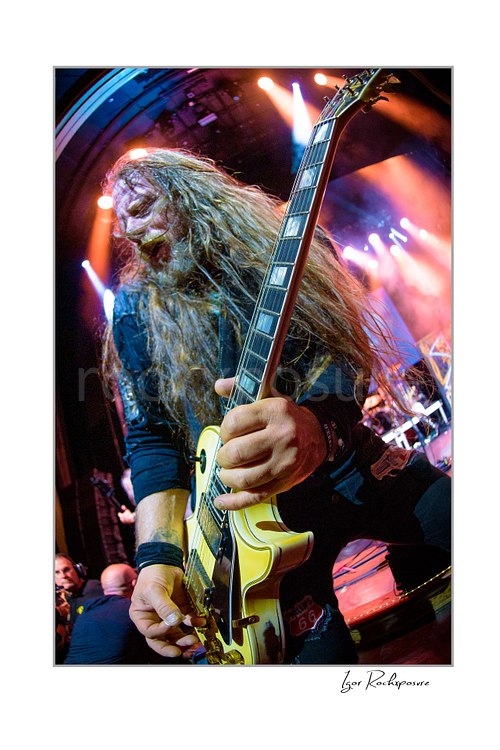 Vertical color image of Chris Latham of Beasto Blanco playing guitar in a dramatic close-up, with long hair in motion and vivid stage lights glowing behind him