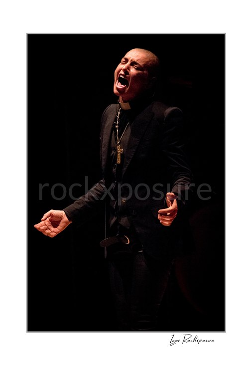 Sinéad O’Connor sings with her mouth open and both hands extended near her waist, wearing a black jacket and cross necklace against a dark background.