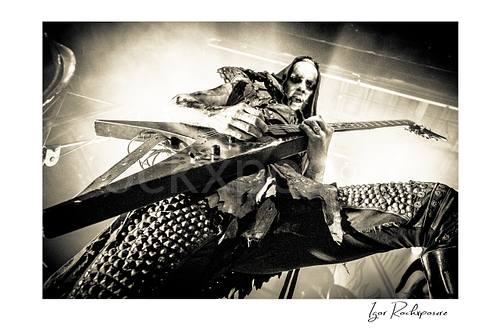 Horizontal black and white image of Nergal of Behemoth playing guitar in an intense low-angle close-up under bright stage backlight