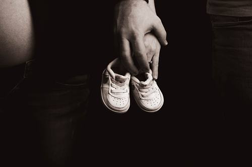 Eine detailreiche Schwarz-Weiß-Aufnahme von zwei sich haltenden Händen eines Paares, die gemeinsam ein Paar kleine, weiße Babyschuhe vor einem dunklen Hintergrund halten.